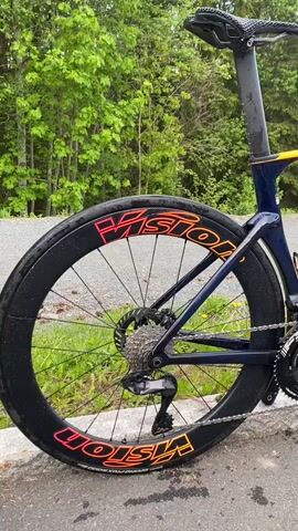 The video shows a Pinarello road bike parked on the edge of an asphalt road. The bike is a sleek design with a dark blue frame that fades into red and orange near the handlebars. It has two black water bottle cages attached to the frame. The wheels are black with the word "Vision" in a bright orange and pink font on the sides. The bike has a black seat and a black chain. The background consists of a green grassy area and a line of trees. The video appears to be shot in a rural setting.