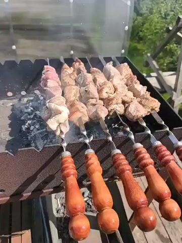 The video shows skewers of meat being cooked on a grill. The grill is filled with charcoal, and the meat is smoking. The skewers have wooden handles and are placed close together on the grill. The meat is a mixture of raw and cooked pieces, with some pieces appearing well-done. The background shows greenery and a clear structure.