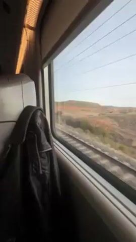 The video shows a view from a train window. Outside, the train is passing by a landscape of dry, reddish earth with sparse vegetation.  Electrical lines are visible along the tracks. Inside the train car, a dark jacket hangs on the back of a seat.  The seat is light colored.  The interior of the train appears modern.