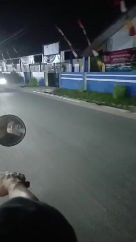 The video begins with a first-person perspective from a moving vehicle, likely a motorcycle, at night. A blurred rider on another motorcycle is visible ahead, illuminated by its headlight. The rider is wearing a red shirt with the number "15" prominently displayed on the back. The background shows buildings with blue and white accents, and red and white flags are strung along a line. The road is paved, and there is some vegetation along the side. The camera continues to record as the vehicles move forward, capturing the motion blur of the passing scenery. The video abruptly ends.