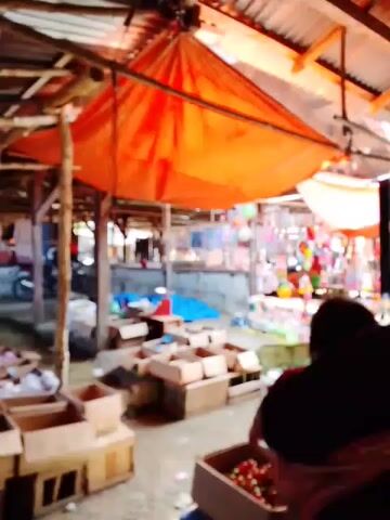 The video shows a market scene from a first-person perspective. The camera moves through a bustling marketplace with stalls displaying various goods. People are seen interacting with the merchandise and each other. The environment appears to be outdoors or in an open-air structure with a corrugated metal roof supported by wooden beams. The stalls are filled with items such as produce, toys, and household goods. The camera focuses on the goods and the people around them, offering a glimpse into the daily life and commerce of the market.
