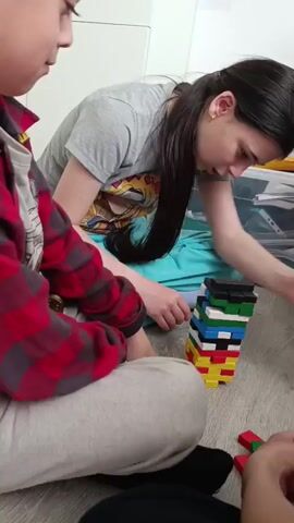 A young boy and woman are playing a game together on the floor. The boy sits in the foreground wearing a red plaid shirt and grey sweatpants. The woman is leaning over a tower of colorful plastic blocks, appearing to try to remove one. She has long dark hair in a ponytail and wears a grey t-shirt and turquoise sweatpants. The floor is light-colored wood, and there are a few loose blocks scattered around the tower.