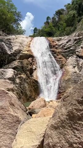 The video shows a waterfall cascading over rocky terrain. The water plunges from a height, landing in a small pool at the base. Large boulders are scattered around the pool and in the foreground. The surrounding landscape is lush with green trees, and a partially cloudy sky is visible overhead.