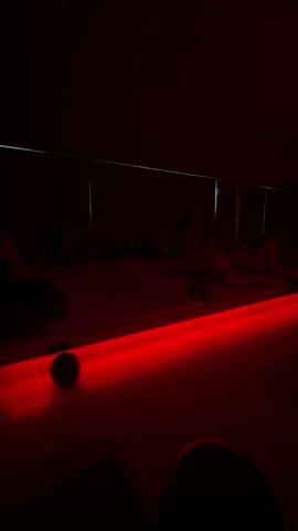 The video shows a dimly lit room with a red light strip extending toward the back.  A line of figures, likely people, are visible but indistinct in the dark interior of the room. A dark, spherical object sits near the front of the red light strip. It is difficult to discern any specific actions or interactions due to the lack of light. The environment appears to be an indoor space, possibly a gym or studio, given the layout and arrangement of the objects and figures.
