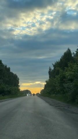 The video shows a POV shot from a vehicle traveling on a wet asphalt road. The road is lined on both sides by dense green trees. In the distance, a sunset can be seen with clouds of yellow, white, and gray. There are a few vehicles on the road ahead, including cars and what appears to be a motorcycle. The sky is overcast, but the sun is breaking through the clouds, casting a warm glow. The overall scene is one of a scenic drive on a cloudy day with a sunset in the distance.