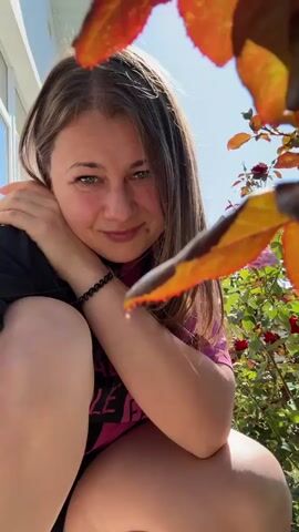 The image features a young woman outdoors, with a bright, sunny sky visible in the background. She is crouching down, looking directly at the camera with a gentle smile. Her hair is long and light brown, framing her face. She is wearing a pink t-shirt and a black bracelet.

In the foreground, a few vibrant orange and red leaves are blurred, suggesting they are close to the camera and possibly falling or swaying. Behind the woman, a rose bush with red and pink roses adds a touch of color to the scene. The woman's posture and the soft lighting suggest a relaxed and pleasant atmosphere.

The overall impression is one of a casual, outdoor moment captured on a sunny day, possibly in a garden or backyard given the presence of the rose bush and leaves. The time of day appears to be daytime, likely mid-morning to afternoon, due to the bright sunlight and the angle of the shadows. There is no visible text in the image.