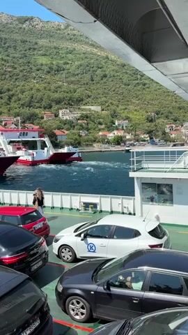 A ferry is docked and cars are parked on its deck. A woman with long dark hair is standing on the ferry's deck and is taking a photo with her phone. In the background, a small town is visible on a hill overlooking the water. The sky is clear and blue, indicating a sunny day. The ferry is white with red accents. There are several cars of different colors parked on the ferry's deck. Some people are standing and walking around on the ferry. The ferry is surrounded by blue water.