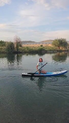 A person is paddleboarding on a body of water, likely a lake or calm river, in Ақбұлақ, Kazakhstan. They are kneeling on the board, holding a paddle and appear to be enjoying a peaceful moment. The sun is setting, casting a warm glow on the scene and creating a serene atmosphere. The sky is partly cloudy, with soft light reflecting on the water. The shoreline is lined with trees and bushes, with rolling hills in the background, suggesting a natural, rural setting. The overall mood is tranquil and relaxed, conveying a sense of leisure and enjoyment of the outdoors. There is no visible text in the media file.