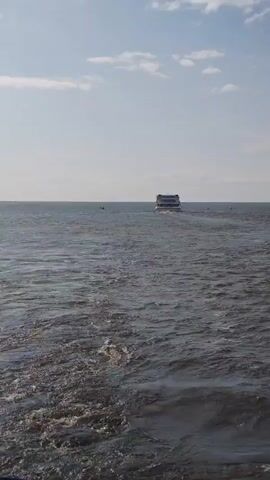 A large boat moves away from the camera across a dark, rippling body of water. The sky above is clear and blue with scattered white clouds. The boat is white with a dark roof and appears to have multiple levels. The water is choppy and creates a wake behind the boat. The horizon is visible in the distance.