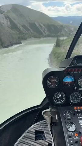 A helicopter flight view from the cockpit of a R44 Clipper II helicopter. The helicopter is flying over a wide, turquoise river that curves through a valley. The riverbanks are lined with trees, and steep, green hills rise on either side of the valley. The sky is partly cloudy, with patches of blue visible. The dashboard of the helicopter is in the foreground, showing various instruments and gauges. The view is from the perspective of the pilot.