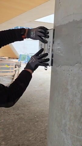 A man wearing a black long-sleeved shirt and black work gloves is using a TriFlex tool to smooth a patch of material on a concrete column. He is pressing the tool against the column with both hands, moving it slightly up and down. The column is under a concrete structure. In the background are wooden pallets, construction materials, and a building with glass windows. The ground appears to be concrete.