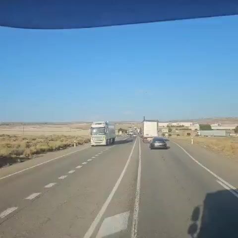 The video shows a traffic jam on a highway. Multiple trucks and cars are stopped on the road, extending as far as the eye can see. The sky is clear and blue, with a few clouds visible in the distance. The landscape on either side of the road appears to be dry and somewhat barren, with rolling hills and sparse vegetation. The sun is shining, casting long shadows on the road. The vehicles are moving very slowly, indicating a significant traffic backup.