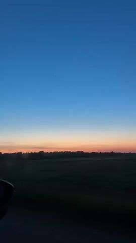 The video, shot from the inside of a moving car, features a landscape during the dawn or dusk. The sky transitions from a deep blue at the top to orange and pink hues near the horizon. Below the sky, a dark strip of trees line the horizon, with a large, dark green field stretching out in front of them. A small portion of the car's side mirror is visible in the lower left corner.  The camera slightly shifts due to the movement of the car. The overall scene is tranquil, capturing the natural beauty of the sky. Music plays in the background.
