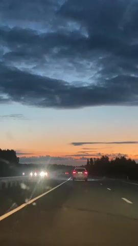 The video shows a view from a car driving on a highway at dusk. The sky is filled with dramatic, dark clouds tinged with the orange and pink hues of a sunset. Several cars are visible on the road, with their headlights on. The trees line the side of the highway, silhouetted against the colorful sky. The road surface appears wet, possibly from recent rain, creating reflections of the headlights.