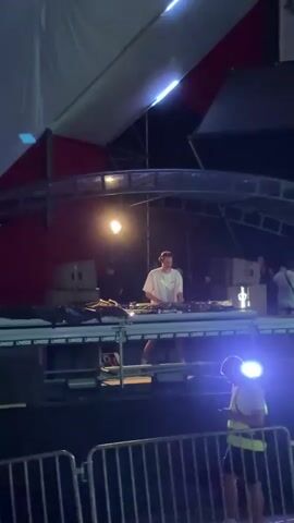 A male DJ in a white t-shirt and dark shorts stands behind a DJ setup on a stage. He is wearing headphones and appears to be mixing music. The stage is lit by spotlights, and there are speakers and other equipment visible. In the foreground, a man in a yellow safety vest and dark shorts is looking at his phone. He is standing behind a metal barricade. The scene appears to be an outdoor event at night.
