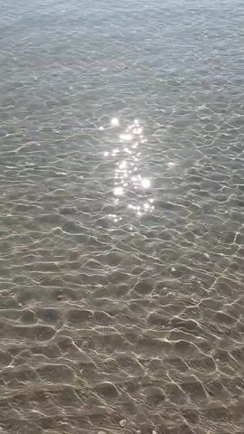 The video shows a shallow body of water with a sandy bottom. Sunlight glints off the surface of the water, creating a shimmering effect. Several small boats are visible in the distance, along with a rocky coastline. The water appears clear and calm, suggesting a peaceful environment. The footage is shot from a low angle, close to the water's surface.