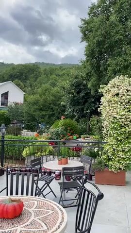 The video showcases a tranquil outdoor patio or terrace with a panoramic view of a lush green, mountainous landscape under a cloudy sky. The camera slowly pans from right to left, revealing the serene setting.

In the foreground, there are two outdoor dining tables with black metal chairs. One table has a distinctive mosaic top with a decorative, reddish-orange pumpkin resting on it. The other table features a few small potted plants. A black wrought-iron railing separates the patio from the garden below. Along the base of this railing, numerous glass jars, filled with what appear to be homemade preserves or jams of various colors, are neatly lined up.

In the mid-ground, a vibrant garden with assorted plants and colorful flowers is visible, behind which a white house with a dark roof and a balcony stands out against the dense green backdrop of trees. A car is partially visible near the house. The entire scene is surrounded by abundant greenery, suggesting a natural and peaceful environment, characteristic of the Meterizi region in Montenegro. The overcast sky indicates a diffused, soft light, possibly late afternoon or early evening. No people are visible, emphasizing the quiet ambiance.