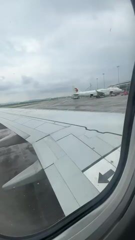 The video is a first-person view from an airplane window. The camera is pointed out the window, showing the wing of the airplane in the foreground. Beyond the wing, an airport tarmac is visible, with several other airplanes parked in the distance. The sky is overcast and cloudy. The camera appears to be stationary.