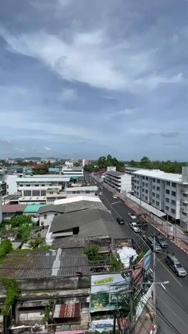 The video is an aerial view of a city street with traffic, buildings, and vegetation under a cloudy sky. The camera is positioned high up, looking down at the scene. In the foreground, there are some older buildings with weathered roofs and overgrown plants. The street is lined with a mix of modern and older buildings, some with balconies and awnings. Cars and a few motorcycles are driving on the street. In the distance, there are more buildings and some hills. The sky is partially covered with white and gray clouds, with patches of blue sky visible. The overall impression is a busy urban environment with signs of development and nature intertwining.