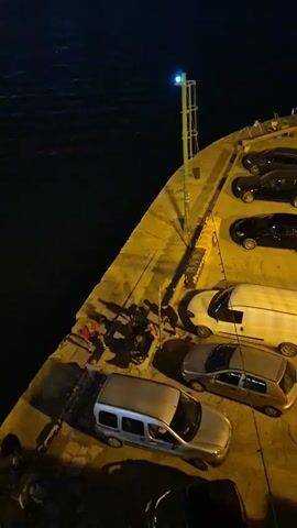 The video shows a nighttime scene of a harbor. Several cars are parked on a concrete pier, illuminated by yellow lighting. In the foreground, there are two silver vans and a silver hatchback. Further back, there are two black SUVs, a white van, and more cars. On the left side of the pier, a group of people are gathered. They appear to be sitting or standing near the edge of the water, with some fishing rods visible. The water is dark, and in the distance, lights from a town or city are visible across the harbor. The overall atmosphere is calm and still.