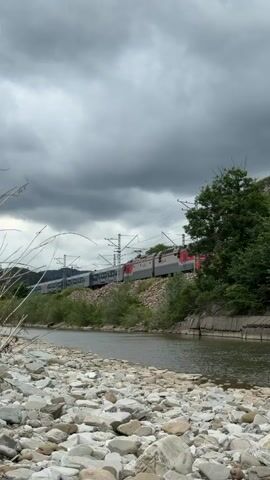 A train moves along a track beside a river on a cloudy day. The train is composed of multiple cars, most of which are dark blue and gray, with some red accents on the front cars. The track is elevated on a rocky embankment. The river is wide and shallow, with a rocky shoreline in the foreground. Lush green trees and vegetation line the riverbank and the embankment where the train tracks are situated. The sky is filled with dark, heavy clouds, suggesting overcast or potentially stormy weather. The scene is shot from a low angle, with the river and rocky shore occupying the bottom portion of the frame.