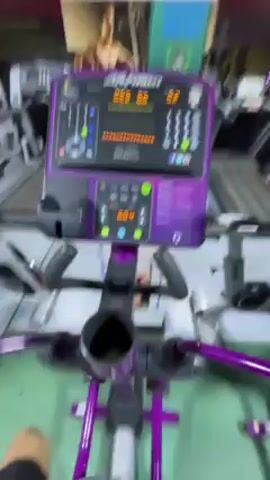 The video shows a person's perspective of using a purple and gray Life Fitness stationary bike in a gym. The bike's console is visible, displaying data, with other gym equipment like treadmills in the background. The camera is mounted on the bike and vibrates as the person pedals.  There is a poster of a shirtless man in the background.  The gym appears to be well-lit.