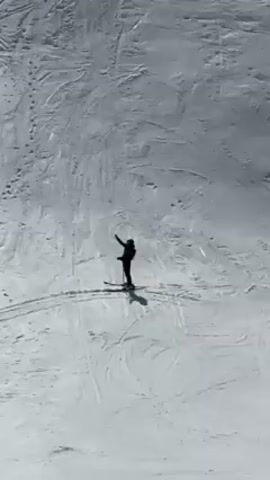 The video shows a snowy mountain landscape with a single skier. The skier is standing still on a slope, waving their arm in the air. The camera then zooms out to reveal a wider view of the mountain range, with several ski slopes visible, and a partly cloudy sky overhead.  The sun is shining through some of the clouds.