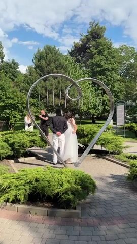The video shows a wedding photo session outdoors in a park-like setting. A couple, the bride and groom, stand embracing within a large, stylized metal heart sculpture. The groom wears a black shirt and white pants, while the bride is in a white wedding dress. They are positioned facing away from the camera, looking towards the left. On the left side of the frame, a person in a black shirt is taking a photo of the couple with a mobile phone. On the right side of the frame, another person is partially visible, also appearing to be taking a photo or filming. The sculpture itself has several metal tubes hanging from the top, resembling wind chimes. The environment is lush with green trees and bushes, and the sky is blue with some clouds. The ground is paved with interlocking bricks. The overall atmosphere suggests a romantic and celebratory occasion.