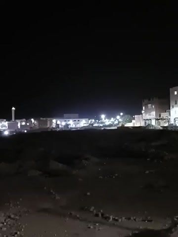 The video shows a night scene of a settlement or town from a distance. The foreground consists of dark, uneven terrain, possibly a field or an area with scattered rocks and dirt. In the background, there are several buildings with illuminated windows and streetlights. One prominent structure appears to be a mosque or religious building with a minaret. The overall atmosphere is dark due to the nighttime setting, and the only sources of light are the artificial lights from the buildings and streetlights. There are no visible people in the foreground. The video is taken from a slightly elevated position.
