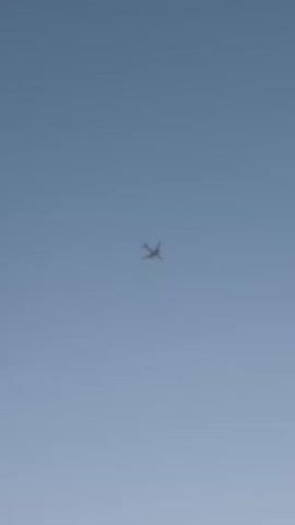 A small, dark silhouette of an airplane is visible against a clear blue sky. The airplane appears to be at a distance and is positioned in the center of the frame. The image is a still frame from a video, indicated by the timestamp in the corner. No other objects or people are visible. The environment is simply the sky.