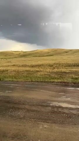 The video opens with a wide shot of a rural, hilly landscape under a cloudy sky. The terrain is covered in dry, yellow grass, suggesting a dry season or arid climate. In the foreground, a muddy, dirt road is visible, with several puddles reflecting the overcast sky. The scene appears to be set in Karagayl, Kazakhstan.

As the video progresses, several vehicles come into view, all on the muddy road. First, an orange truck, possibly a utility or service vehicle, drives by. It is followed by another truck carrying a large, red cylindrical tank. Finally, a bright orange crane truck appears, its boom extended forward. The crane has a hook attached. Text is visible on the side of the crane truck, reading "УЛЬЯНОВЕЦ" (ULYANOVETS). The weather appears to be overcast, possibly indicating rain has recently fallen or is about to. The overall mood is somewhat bleak due to the weather and the utilitarian nature of the vehicles, set against a backdrop of a vast, natural landscape.