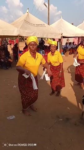 A group of women and two children are singing and dancing outside under white tents, possibly at an event or celebration. The women are dressed in matching yellow shirts, head wraps, and red and brown patterned skirts. Most of them are standing and singing, while one woman is seated, possibly leading the singing. One child is seated in a blue chair, playing what looks like a small drum or percussion instrument. The ground is dusty and there are chairs and other objects scattered around. The weather appears sunny. The video is shot from a slightly low angle.