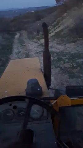 The video shows the perspective from inside the cab of a yellow tractor moving along a dirt path. The sun appears to be setting, leaving the environment dimly lit.  The path is narrow, winding up a slight hill.  The tractor's dashboard and steering wheel are visible. A tall exhaust pipe is next to the driver's side of the cab.  Vegetation is visible along the sides of the path.