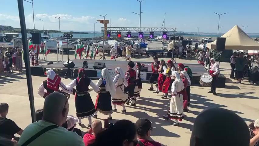 A group of people in traditional Bulgarian folk costumes are performing a dance in a circular formation on a paved outdoor area. They are holding hands and moving in unison. In the background, there is a stage with musical equipment and more people, some of whom appear to be performers or organizers. Several boats are docked in the water behind the stage, and a clear blue sky is visible overhead. The event seems to be a cultural celebration taking place near a harbor or marina.
