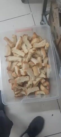The video shows a close-up, top-down view of a clear plastic storage container filled with numerous pieces of cut bread. The bread pieces appear to be of various shapes and sizes, mostly rectangular or elongated, with some appearing to be crusts. The bread is a light brown color, suggesting it may be slightly stale or toasted. The container is placed on a tiled floor, and a portion of a dark-colored shoe is visible in the bottom left corner of the frame. In the background, to the right, there are stacked boxes, one of which has green and white labeling. The lighting in the scene is somewhat dim. The video consists of a series of still frames that create a slight motion effect, as if the camera is subtly moving or zooming in on the contents of the container.