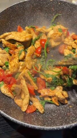 This is a close-up, over-the-shoulder shot of a person cooking chicken, cherry tomatoes, and spinach in a frying pan. The food is being stirred with black tongs. The pan is dark gray and speckled, with steam rising from the food. The background is blurred, but appears to be a kitchen setting, possibly a stovetop. The lighting suggests it is daytime. There is no visible text in the media. The activity is cooking, likely for a meal. The focus is on the food and the action of cooking it.