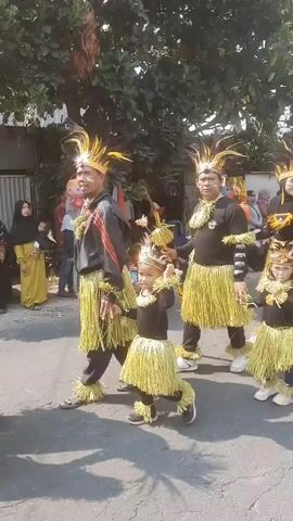 A parade is taking place outdoors on a paved road lined with trees. A group of people, including adults and children, are walking in the parade, all wearing matching costumes. The costumes consist of black tops with yellow fringe skirts and headpieces adorned with yellow feathers. Some individuals are holding staffs decorated with yellow fringe. In the background, onlookers can be seen standing along the side of the road, and there are buildings and more trees visible. The weather appears to be sunny.