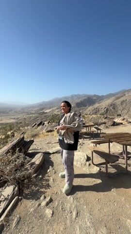 The video shows a person standing on a rocky overlook with picnic tables, smiling and holding a jacket. The camera pans to show a view of mountains and a city in the distance. The landscape is dry and desert-like with scrub brush and scattered rocks. The sky is clear and blue.