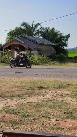 The video shows a rural road with a small house on the left and a field on the right. A motorcycle drives past the house. Further down the road, a yellow dump truck drives by, kicking up dust. A car then passes, followed by another motorcycle. The scene appears to be in a warm climate with clear skies.
