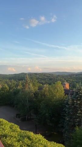 This is a vertical video of a natural landscape at sunset. In the foreground, the tops of green trees are visible. Beyond the trees, there are buildings with brown roofs and chimneys. The middle ground consists of a vast expanse of green trees. In the background, rolling hills are visible against a bright orange and yellow sunset sky with some clouds. The sun is a bright orb, casting a warm glow over the scene and reflecting on a body of water. The overall impression is a peaceful, scenic view of nature and a small settlement bathed in the warm light of the setting sun.