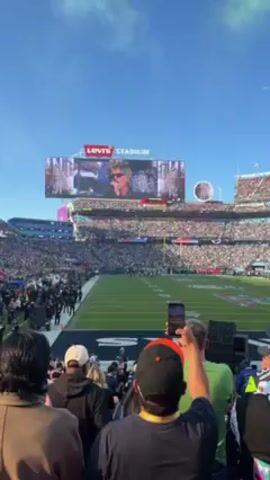 The video shows a view from the stands at Levi's Stadium, filled with a large crowd. A football field is visible in the foreground. On the stadium's jumbotron, an image of a person is displayed. Some people in the foreground are taking pictures or videos with their phones. The sky is blue with some clouds. There are stadium lights and other equipment visible on the field.