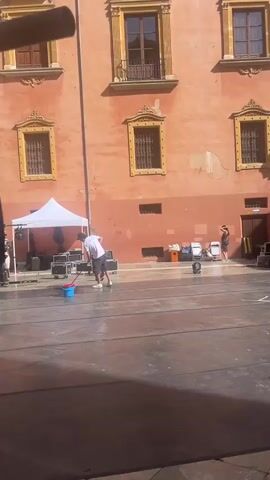 The video shows an outdoor urban scene with a man in a white t-shirt and shorts mopping a large, dark-colored surface, possibly a stage, with a blue bucket and a mop. Behind him, a building with a peach-colored facade and ornate windows is visible. Further back, a woman in a dark dress stands near some equipment. To the right, another building with yellow and orange facades and balconies lines a street where several people are walking. Various pieces of equipment, including speakers and lighting rigs, are set up on the stage area. The overall impression is of a public space being prepared for an event.