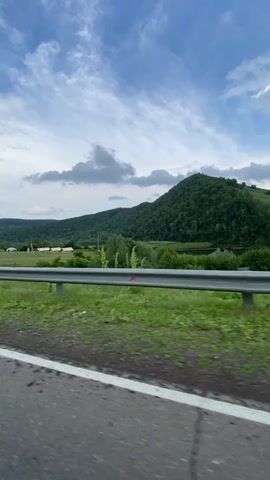 The video shows a view from the inside of a moving car. The camera is positioned to capture the road ahead, the side of the car, and the passing scenery. The scenery consists of rolling green hills covered in trees, a wide-open field with a few buildings in the distance, and a cloudy blue sky. The road is paved with white lane markers. A metal guardrail runs along the side of the road. Reflections are visible in the car's side mirror, including what appears to be a person's hand and arm. The video is shot from a first-person perspective, suggesting the viewer is a passenger in the car.