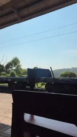 A freight train car slowly moves along tracks outside a covered train station. The freight car appears to be flat. Trees and foliage are visible in the background. The sky is clear and blue. A dark bench sits in the foreground, partially obscuring the view.