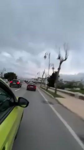 From the perspective of a passenger in a bright yellow or green car, the video shows a road with other cars ahead under an overcast, grey sky. The car is on the right lane of a two-lane road, and the sidewalk to the right has grass and streetlights reminiscent of old-fashioned gas lamps. White buildings are intermittently visible beyond the trees along the sidewalk. There are no visible people. The focus is on the road and the cars in front. The video appears to be taken during daylight hours, but the sky indicates it's a cloudy day.