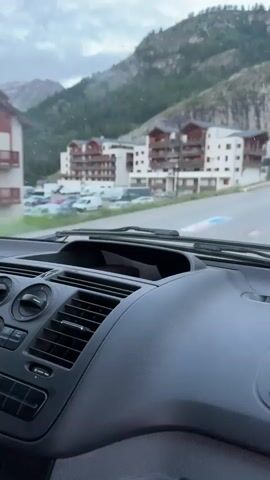 The video begins from the perspective of a passenger inside a vehicle, showing the dashboard and the road ahead. The vehicle is moving along a paved road that curves to the right. On the left side of the road are buildings that appear to be part of a ski resort, with multiple stories and balconies. Behind the buildings are steep, rocky mountains covered with trees. The sky is overcast. As the video progresses, the vehicle passes a white minivan. The road continues to wind through the mountainous terrain, with more buildings and ski lifts visible in the distance. The perspective remains inside the vehicle throughout the short clip.