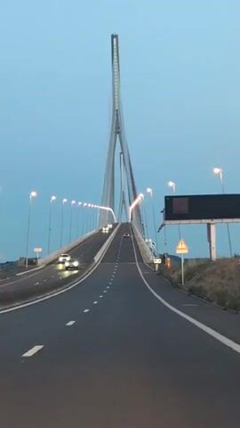 A vertical, low-angle shot shows a wide, empty highway leading towards a tall, modern suspension bridge. The sky is a pale, clear blue, and it appears to be dusk. Streetlights line the highway, and a few cars are visible in the distance, their headlights on. A large digital billboard is mounted on a pole to the right of the highway, and a triangular warning sign with a red border and a symbol of a person walking is visible below it. A red and white windsock is also visible on a pole to the right of the highway. The camera is positioned inside a vehicle, with the edge of the dashboard visible at the bottom of the frame. The overall impression is one of a quiet, uneventful drive on a modern infrastructure project.