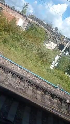 This is a first-person perspective video taken from a moving train. The train tracks are visible in the foreground. In the background, there is a lush green embankment with trees and bushes. Beyond the greenery, there is an old, dilapidated building with a white facade. Some windows are broken, and the roof appears to be in disrepair. Overhead, there are power lines and a blue sky with scattered white clouds. The scene suggests an industrial or abandoned area next to a railway line.