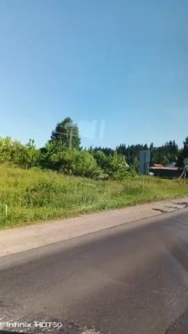 The video shows a view from inside a vehicle looking out at a rural landscape. The sky is clear and blue. On the left side of the road, there is a grassy verge with some bushes and trees. A blue road sign is visible on a pole. Further into the distance, there are houses with roofs of various colors, surrounded by vegetation and trees. On the right side of the road, there is a dirt track and a wooden utility pole with wires. The road itself appears to be paved, with some cracks and uneven surfaces visible. The overall impression is of a sunny day in a countryside setting.