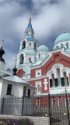The video shows a panning shot of a large, ornate church with red walls and turquoise domes against a cloudy sky. The church features white trim, crosses, and arched windows. A tall bell tower with a blue spire is visible on the left side. In the foreground, a black wrought-iron fence with decorative scrollwork encloses a garden area with low-lying plants. The ground is paved with cobblestones.